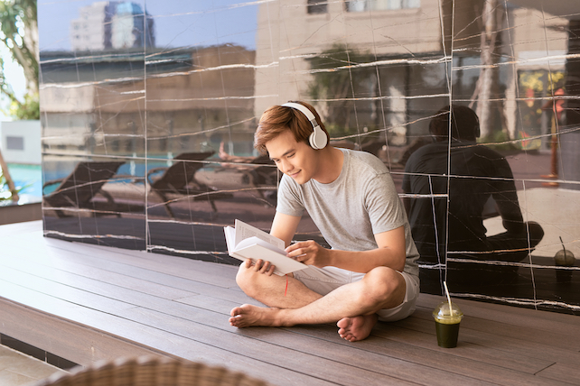 junger Mann mit Kopfhörers sitzt auf einer Terrasse und liest ein Buch