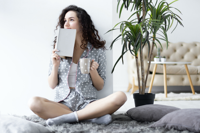 junge Frau im Pyjama sitzt im Schneidersitz auf dem Boden ihres Schlafzimmers und hat einen Kaffee und ein Buch in der Hand - Morgenroutine