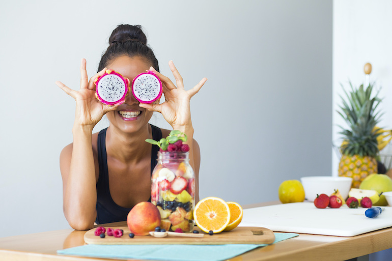 gesund Leben durch Ernährung, Bewegung und Erholung - junge Frau sitzt am Esstisch, vor ihr liegt Obst und sie hält sich Drachenfrucht vor die Augen
