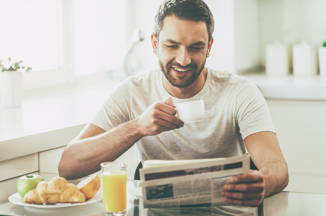 junger bärtiger Mann sitzt am Frühstückstisch und liest die Morgenzeitung - Morgenroutine